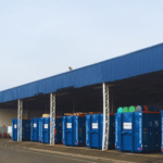 Foto de um galpão industrial com caçambas azuis rotuladas para diferentes tipos de resíduos, como 'Sucata Metálica' e 'Madeira'. Telhado azul e paredes brancas do galpão, com extintores de incêndio visíveis.