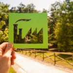 Mão segurando um recorte de papel verde em forma de fábrica, com uma paisagem de árvores ao fundo, representando a transição para práticas industriais mais sustentáveis e ecológicas.