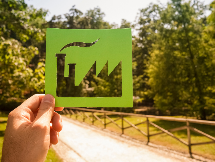 Mão segurando um recorte de papel verde em forma de fábrica, com uma paisagem de árvores ao fundo, representando a transição para práticas industriais mais sustentáveis e ecológicas.