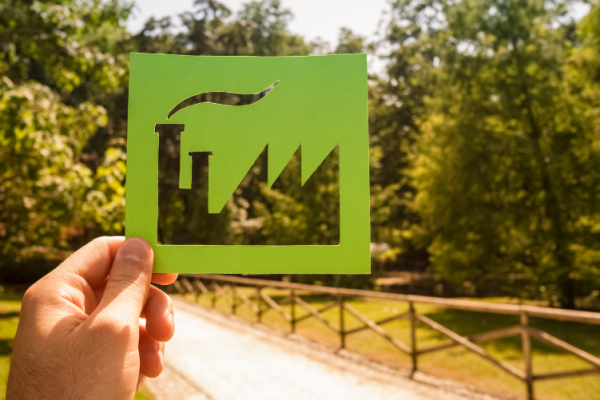 Mão segurando um recorte de papel verde em forma de fábrica, com uma paisagem de árvores ao fundo, representando a transição para práticas industriais mais sustentáveis e ecológicas.